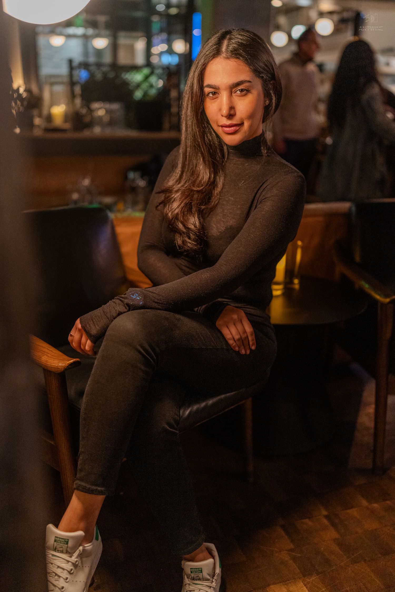 A young brunette woman in a black turtleneck and jeans sits in a chair at a warmly lit restaurant interior.