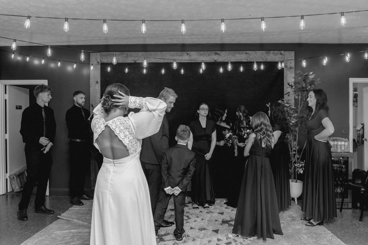 Bride and groom share first dance surrounded by wedding guests under string lights at indoor reception.