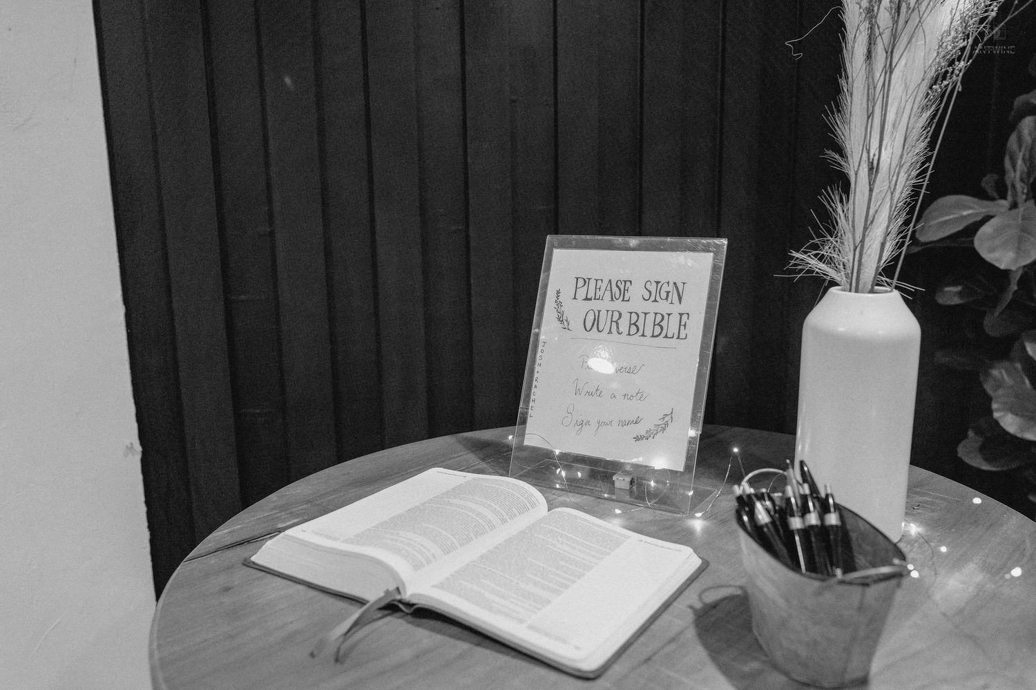 Black and white photo of an open guest book on a round table with a sign, vase, and pen holder at a wedding.