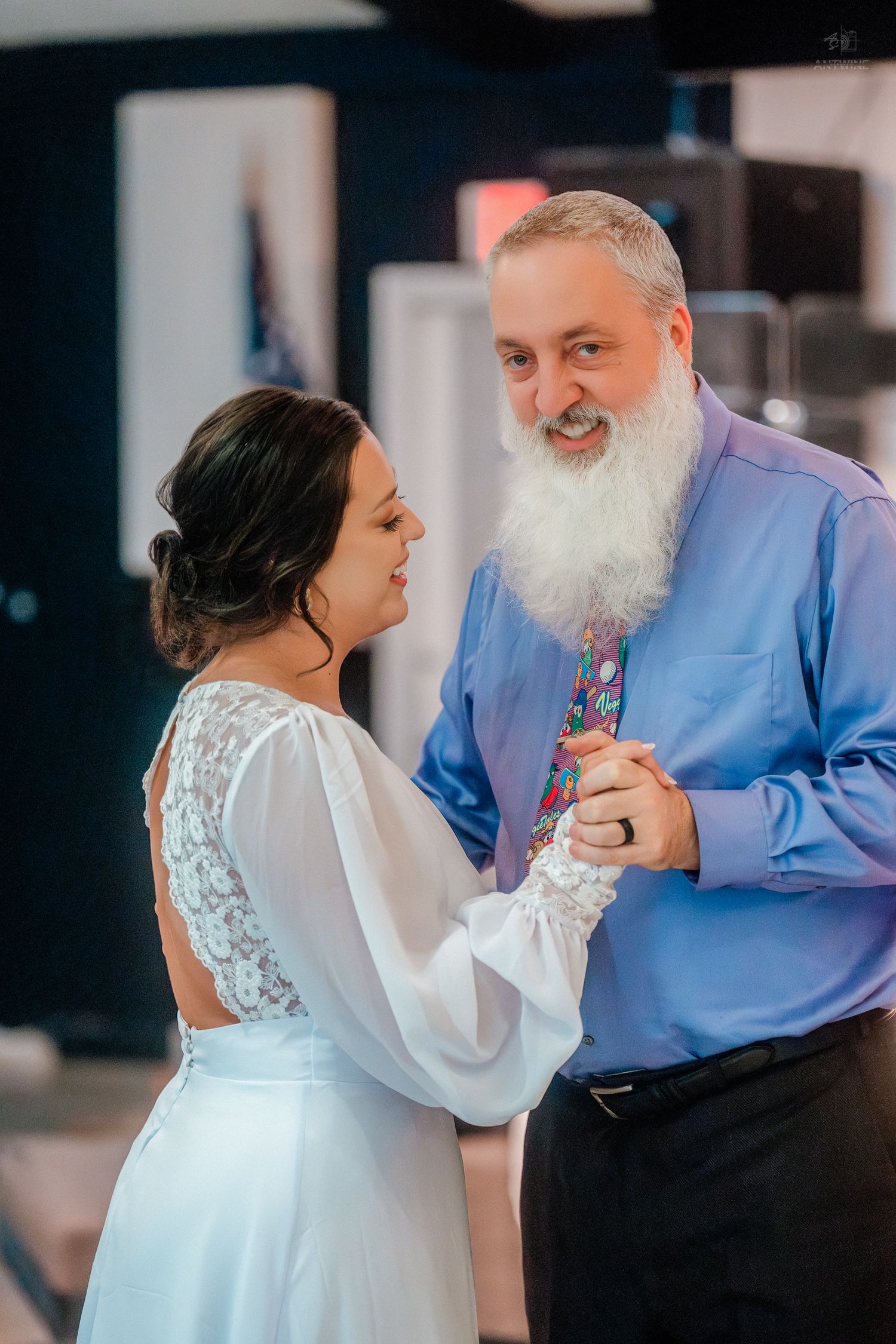 Bride and older bearded man share a dance at wedding reception, smiling warmly at each other.