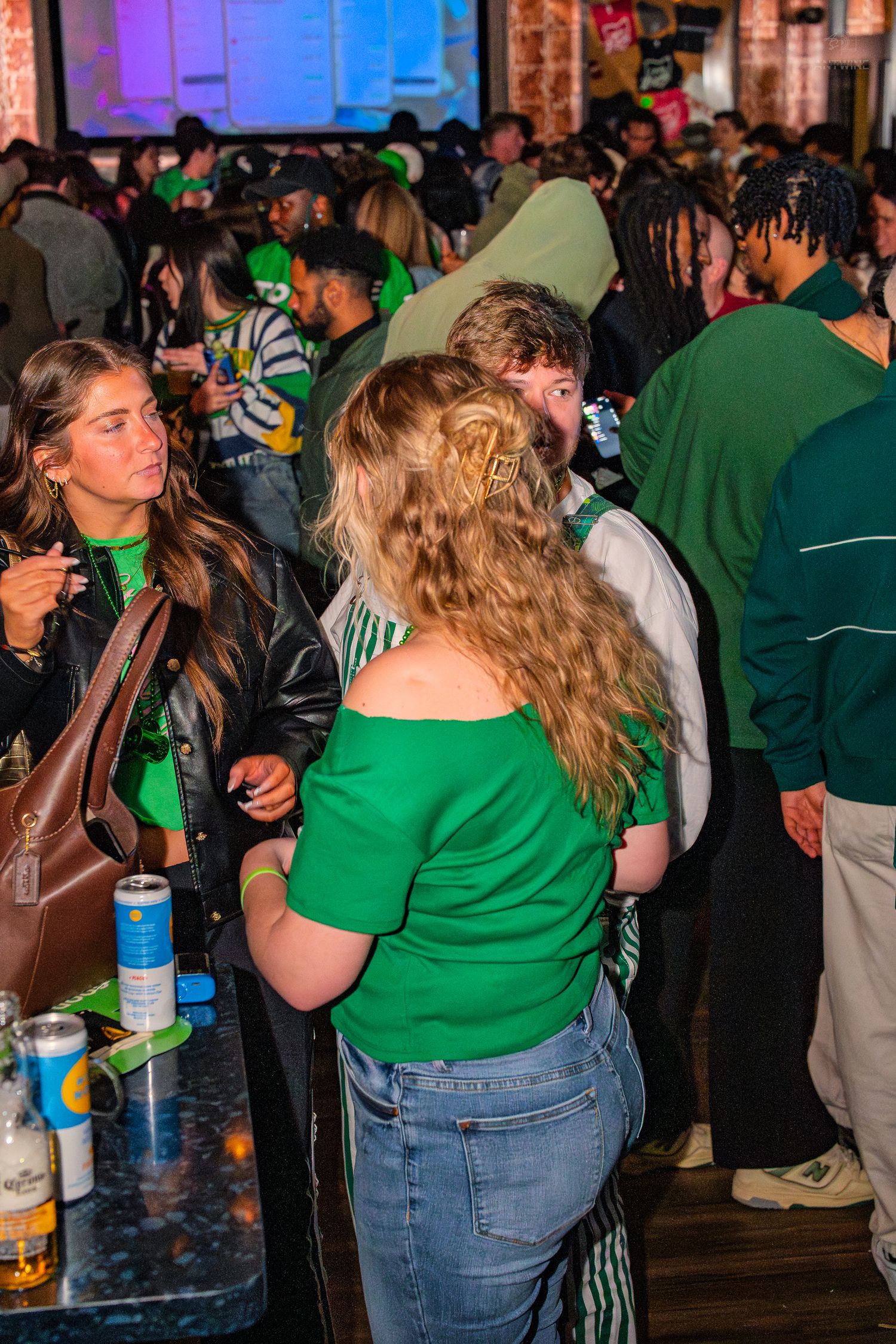 Crowd of young people in green outfits mingling at a busy St. Patrick's Day bar celebration.