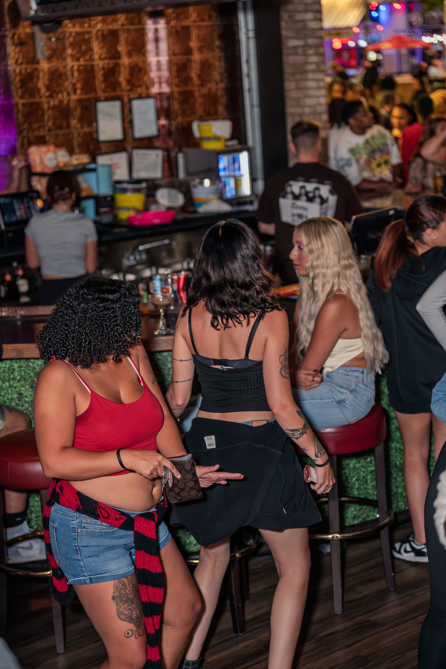 Three women standing at a bar counter, wearing casual outfits with visible tattoos at a nightclub.
