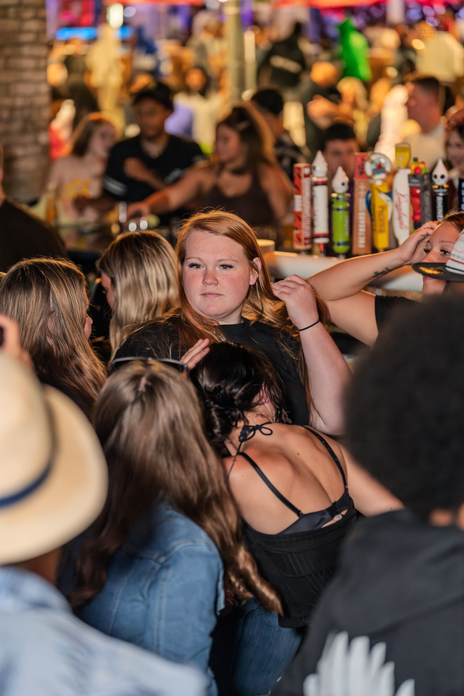 Woman in black shirt stands out in a crowded bar scene with colorful beer taps and patrons visible behind her.