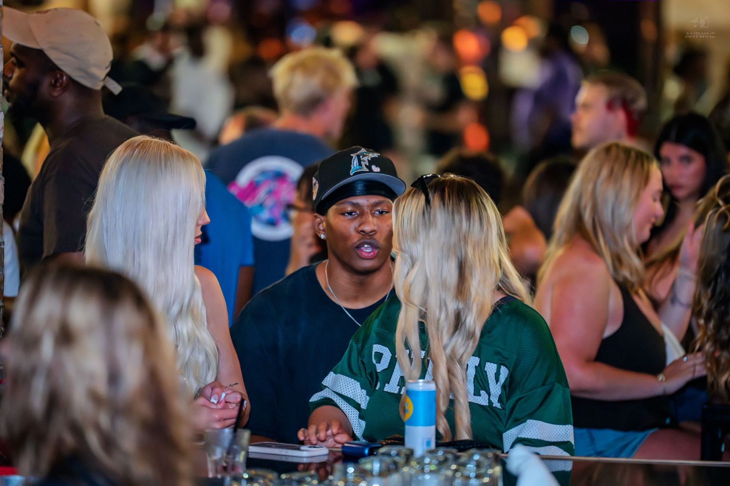 Man in black cap and green jersey talking with blonde women in a busy nightclub.