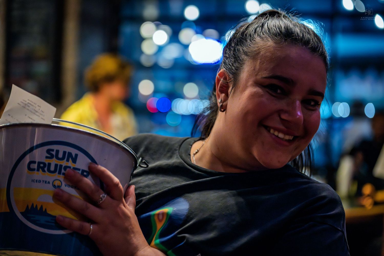 Smiling woman holding a Sun Cruiser drink bucket at a vibrant nightlife venue.