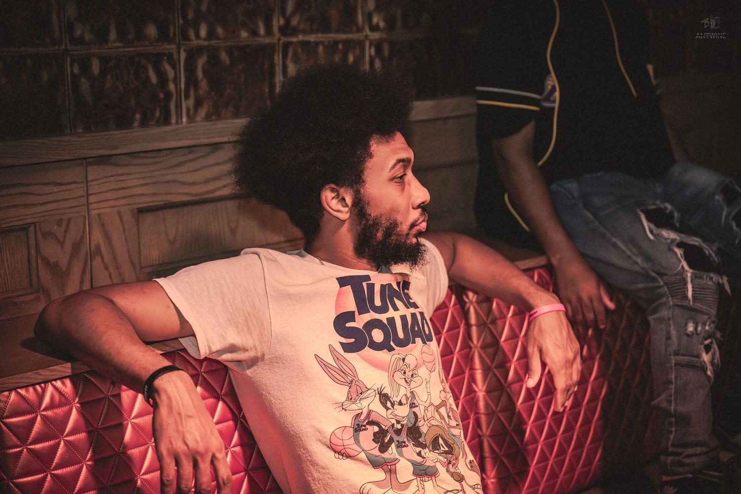 Man in Tune Squad t-shirt relaxing on pink quilted sofa in dimly lit venue.