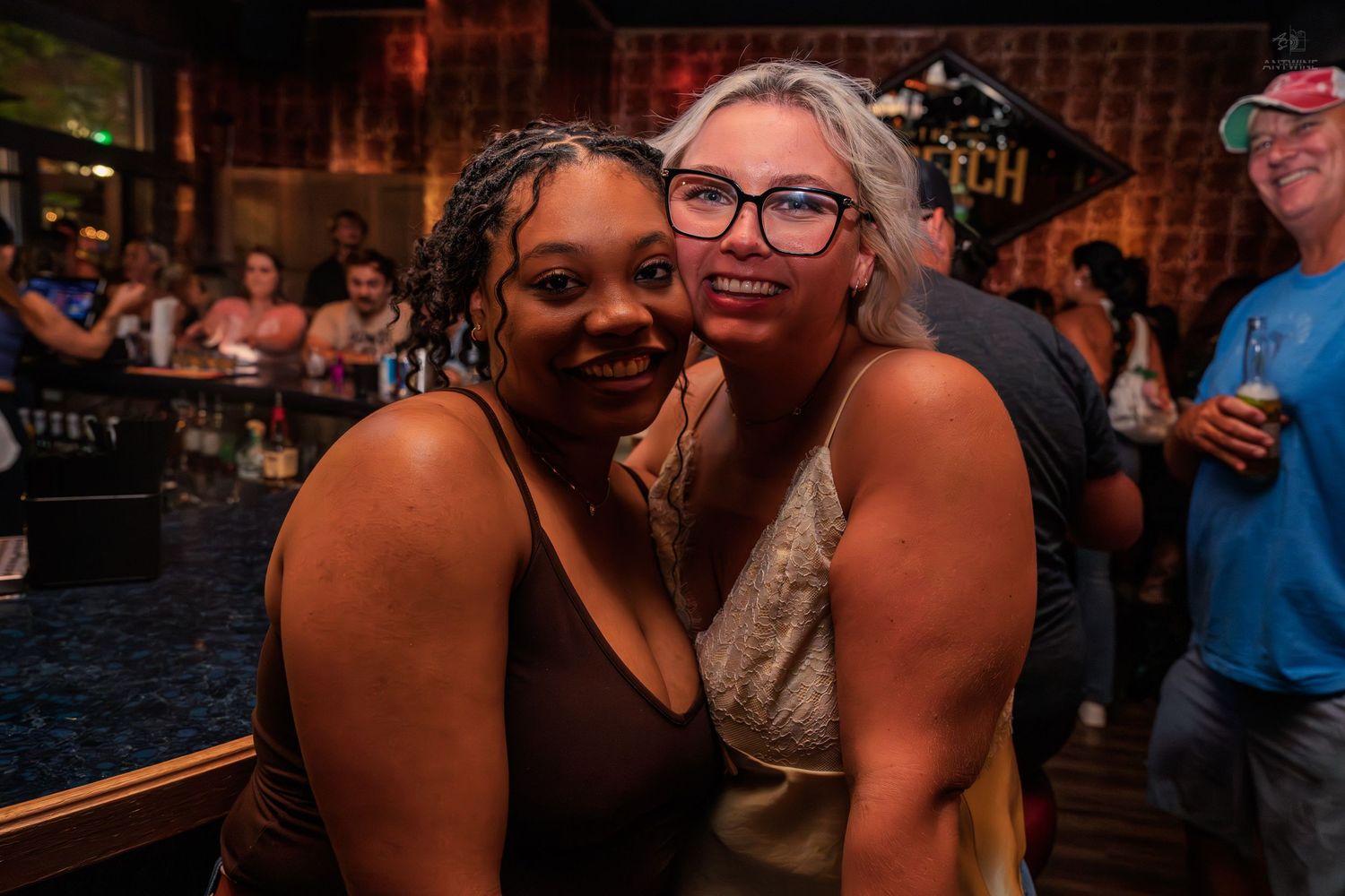 Two smiling women embracing for a photo inside a warmly lit bar at night.