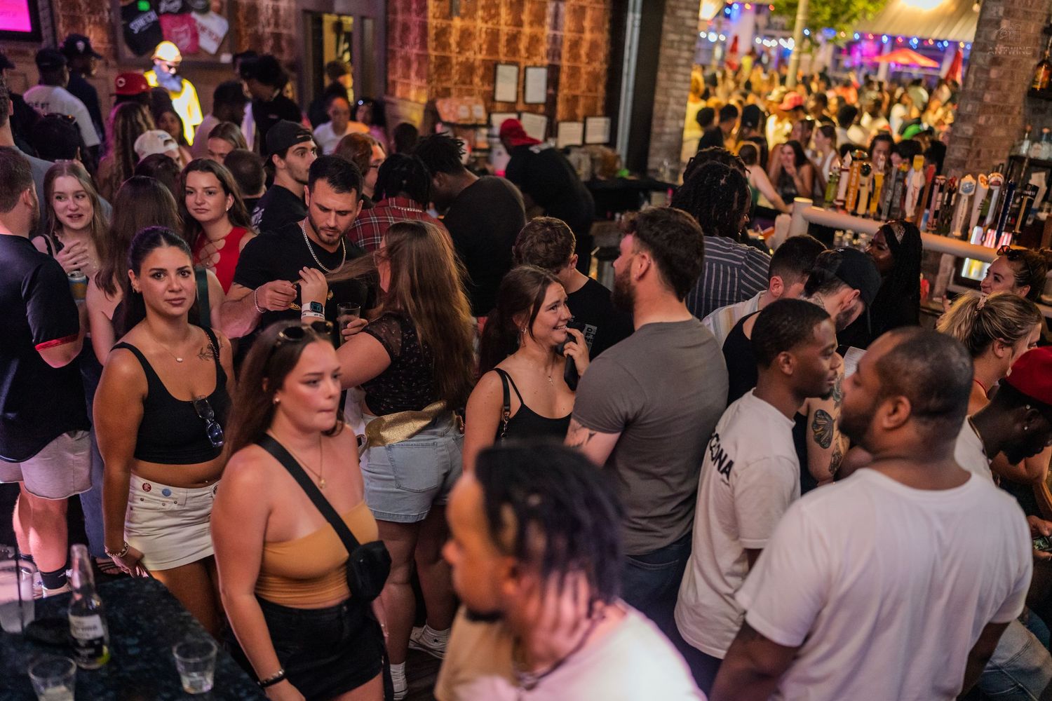 Packed nightclub with crowd of young people dancing and socializing under pink and purple lights.