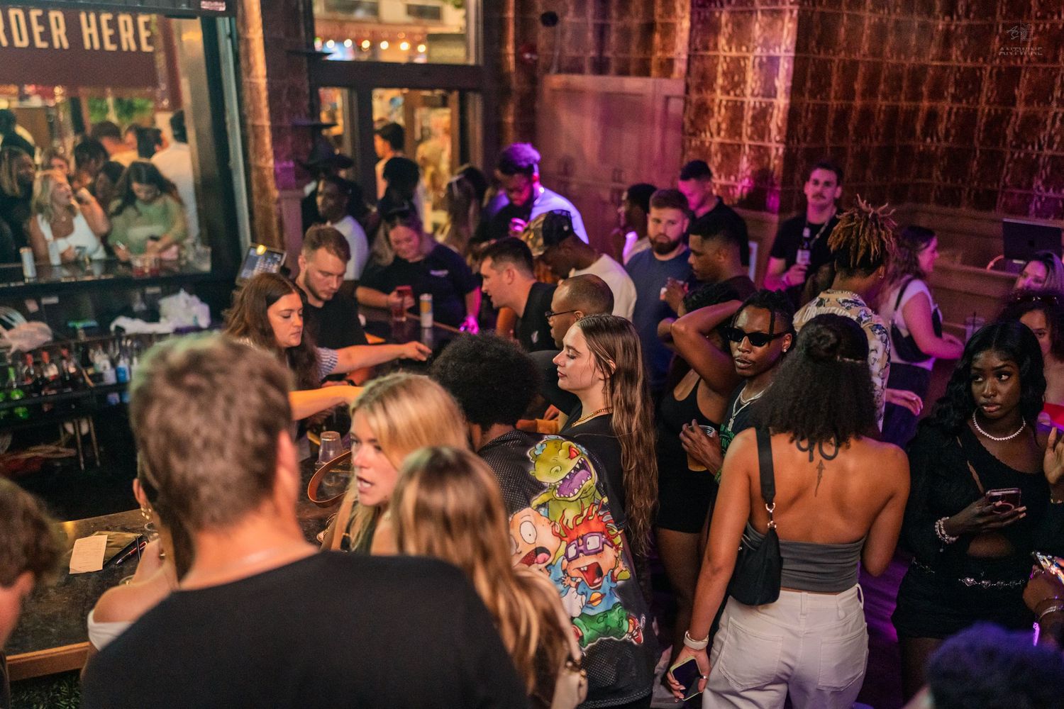 Energetic nightclub crowd mingling outdoors near a bar with colorful purple lighting.
