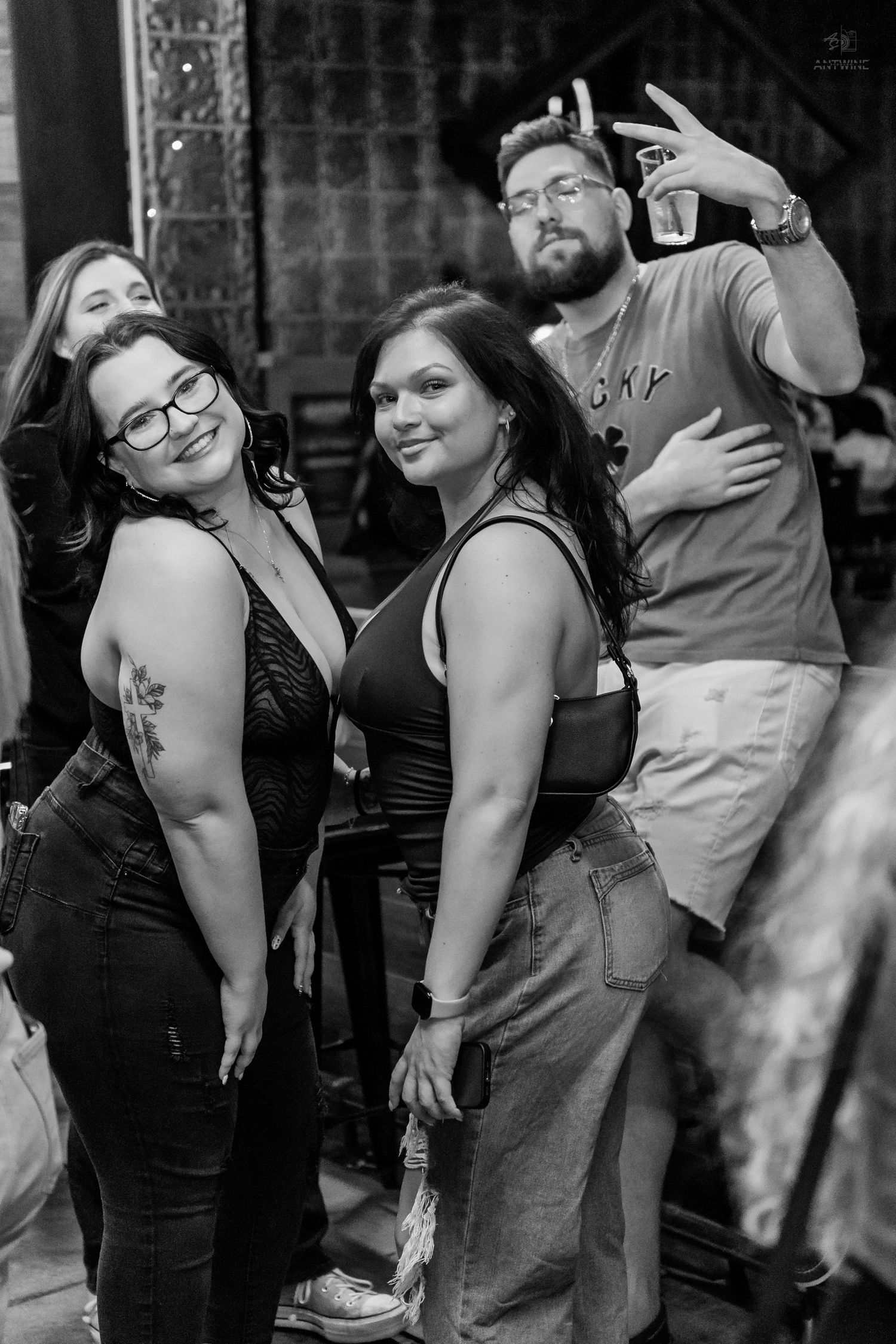 Group of friends posing and smiling at a nightclub, man in background giving peace sign.