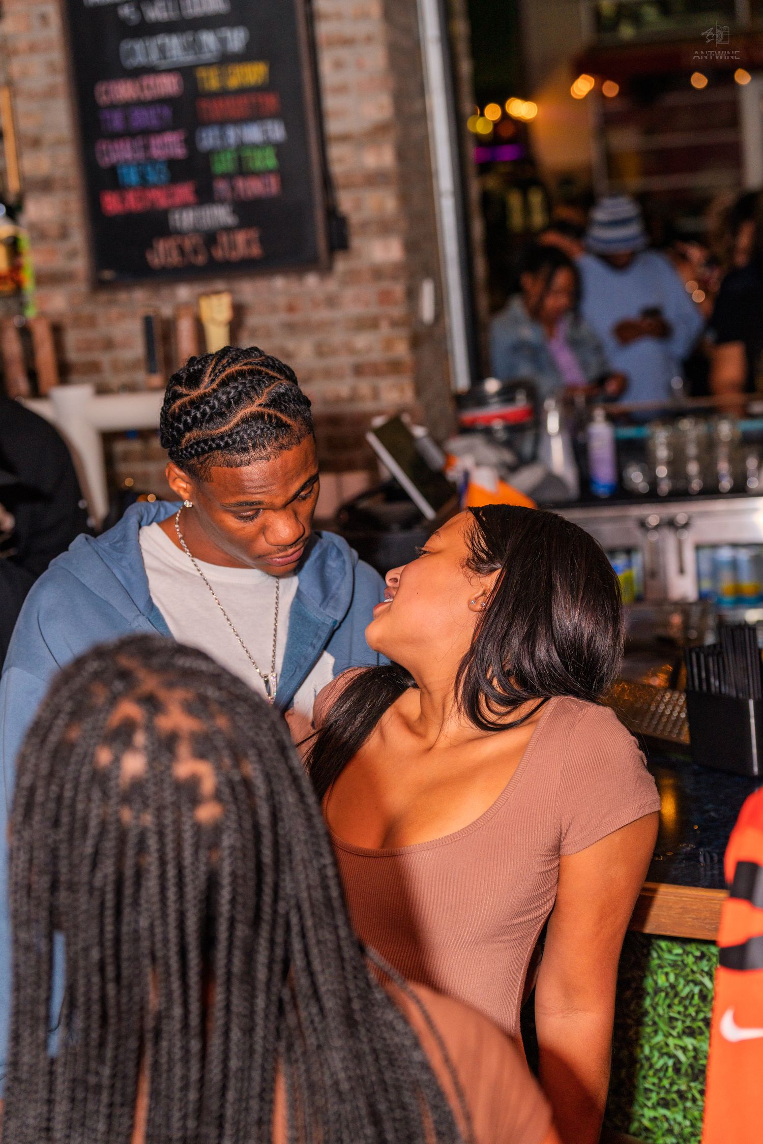 Man and woman laughing together at a bar with cocktail menu chalkboard behind them.