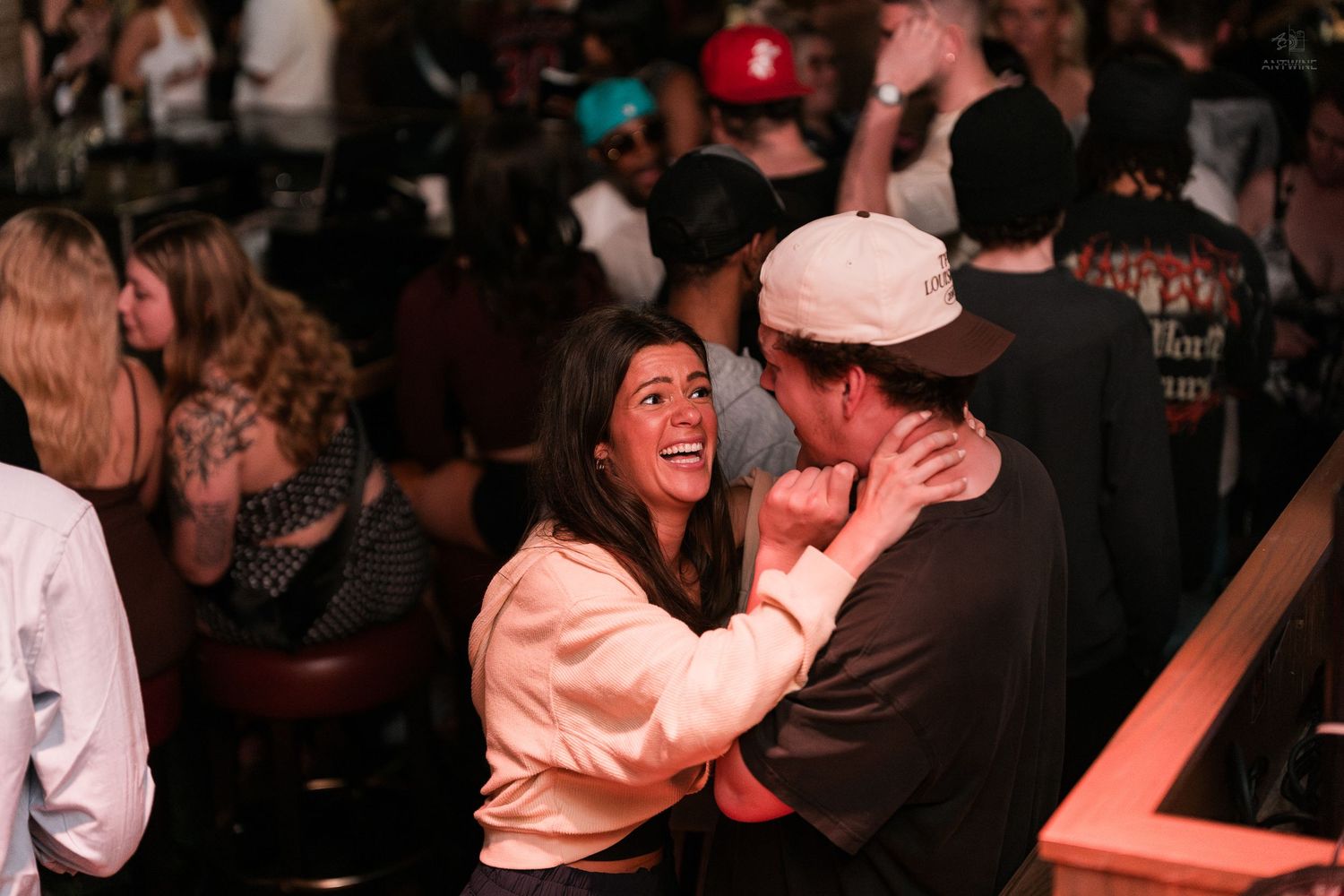 A laughing woman in a beige top shares a joyful moment with a man at a lively bar or nightclub.