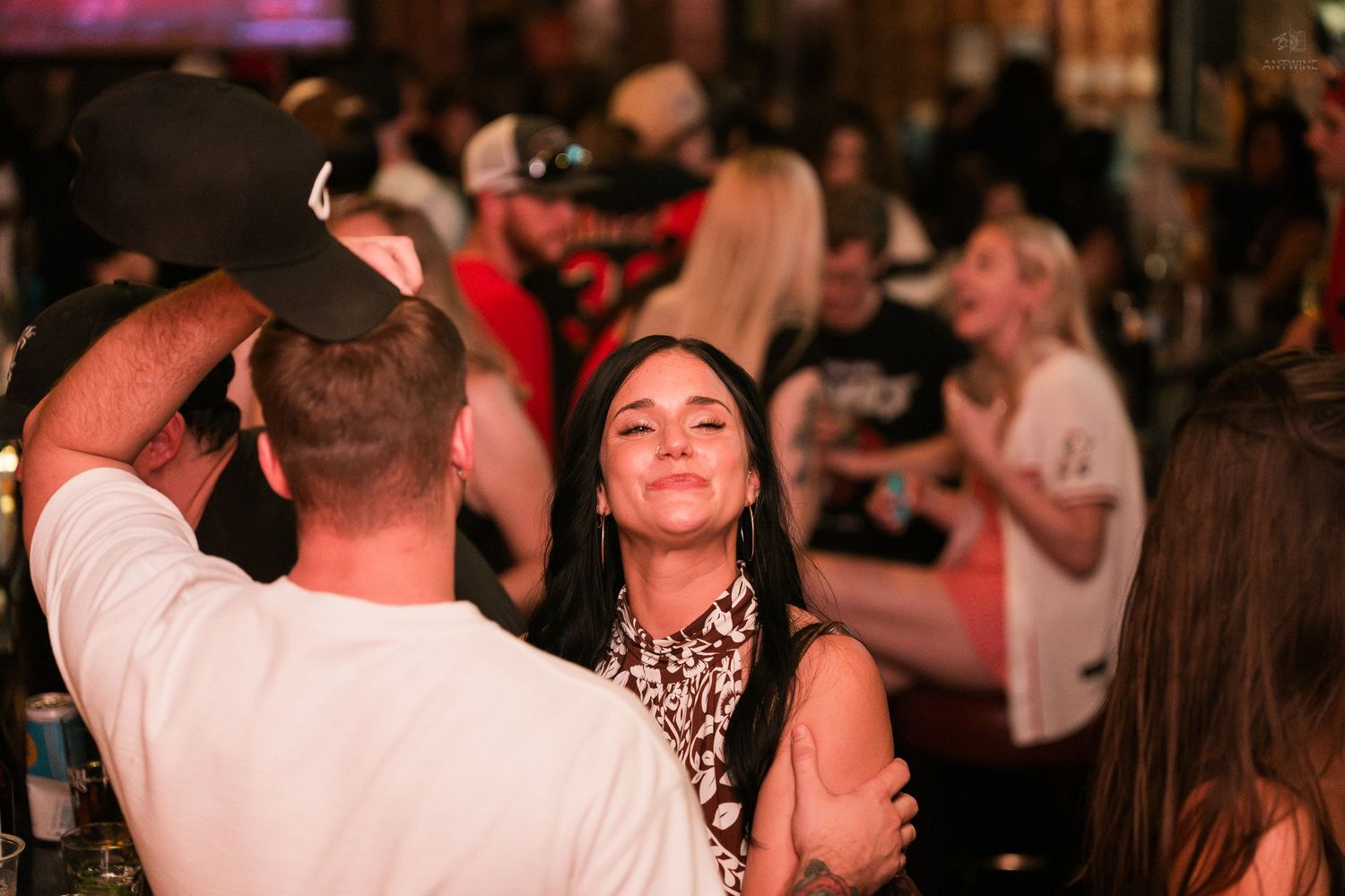 Young woman smiling while dancing with a man in a white shirt at a lively crowded bar or nightclub.