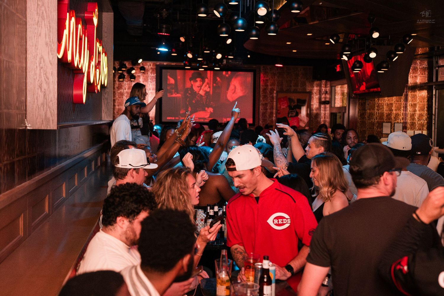 Energetic crowd in Cincinnati Reds gear cheers while watching game at packed sports bar.