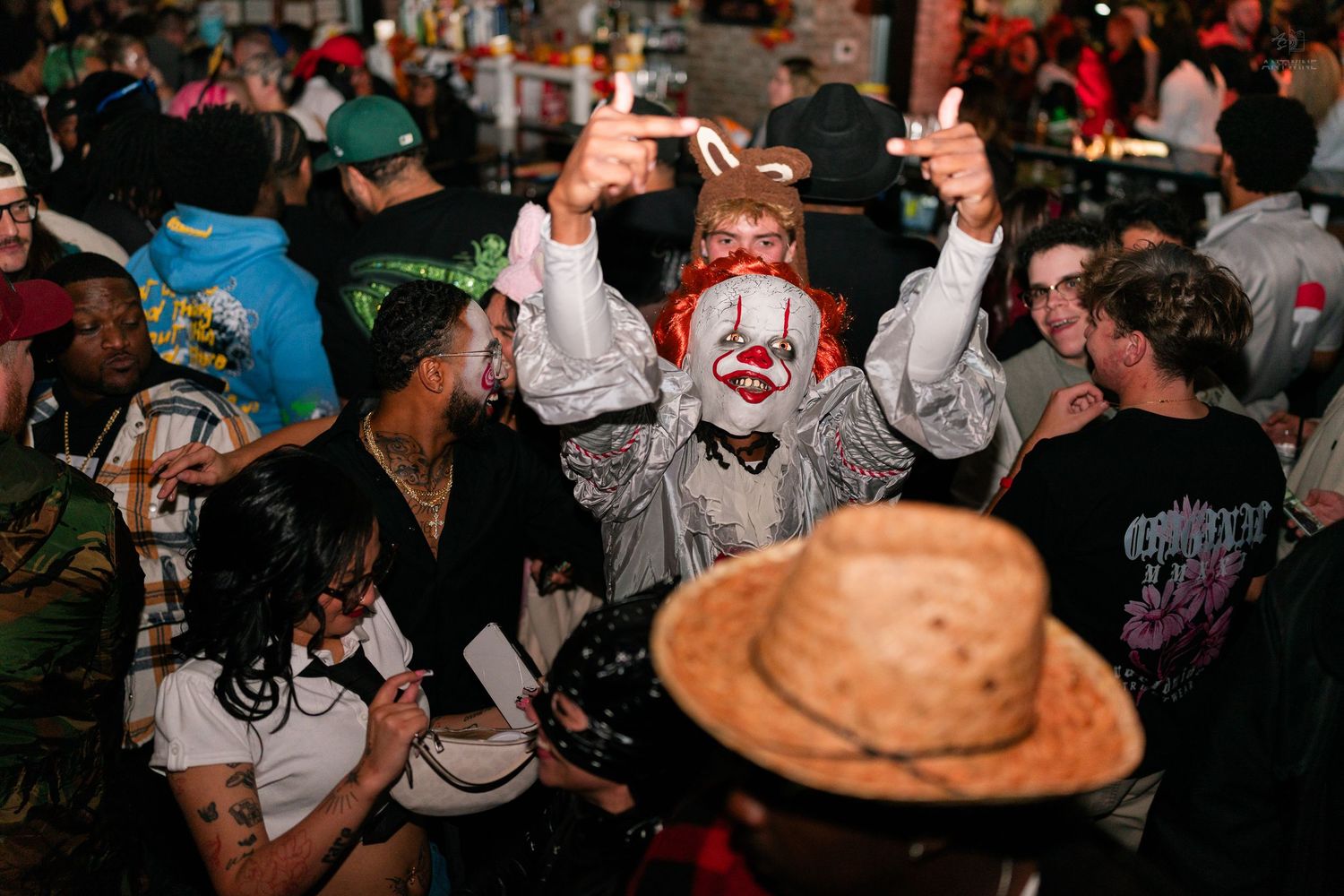 Person in clown costume with face paint raises hands at a crowded Halloween party in a bar.