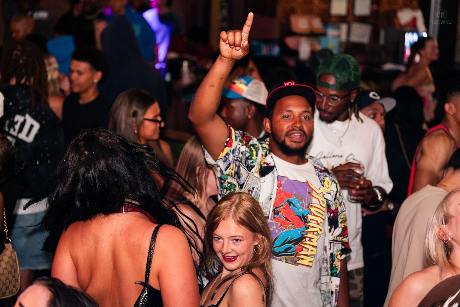Man in patterned shirt dancing with one finger raised at a lively nightclub party.