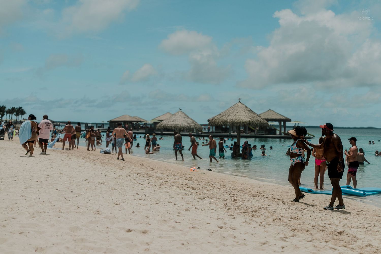 Crowds of tourists enjoy a tropical beach with overwater bungalows and clear blue water.