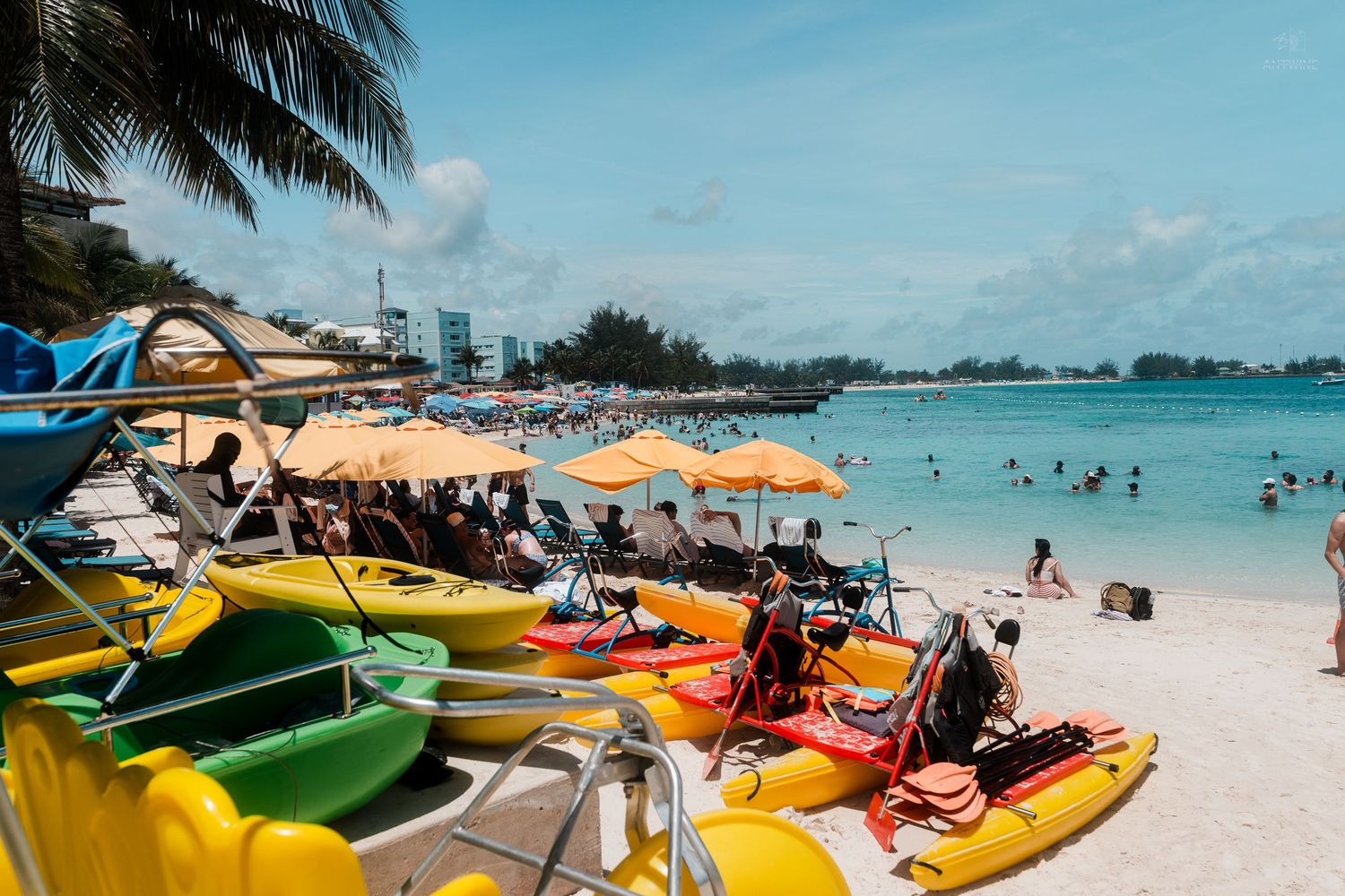 Colorful kayaks lined up on a sunny Caribbean beach with swimmers and umbrellas in background.