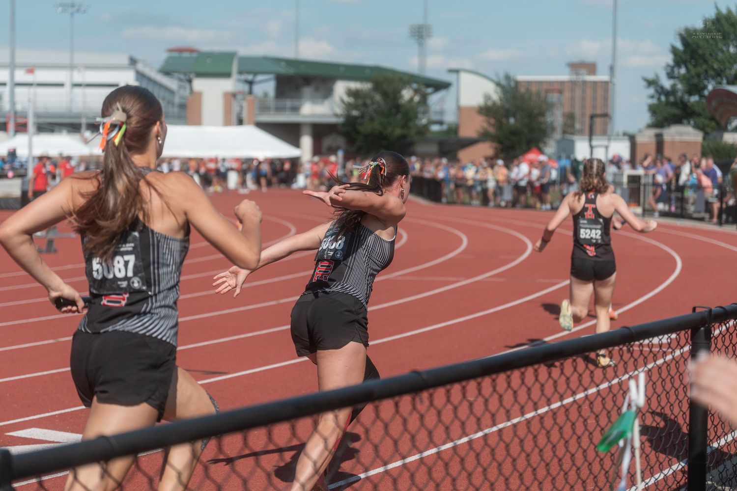 Female relay runners in black uniforms pass a baton during an outdoor track and field competition.