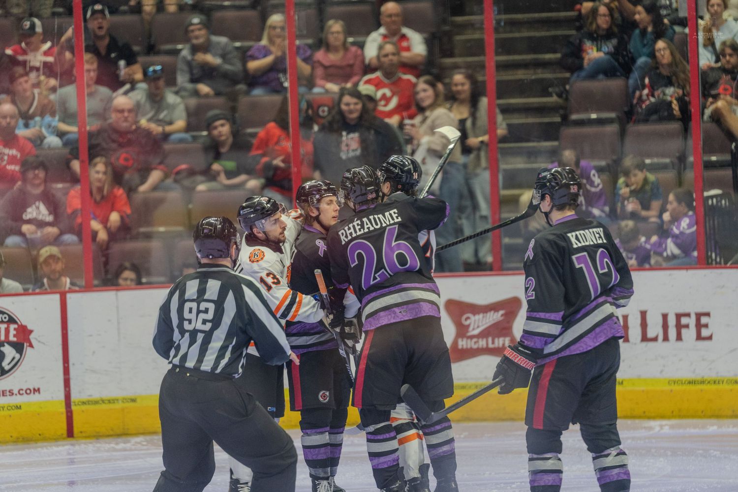 Ice hockey players in purple jerseys crowd around an opponent near the boards during a professional hockey game.