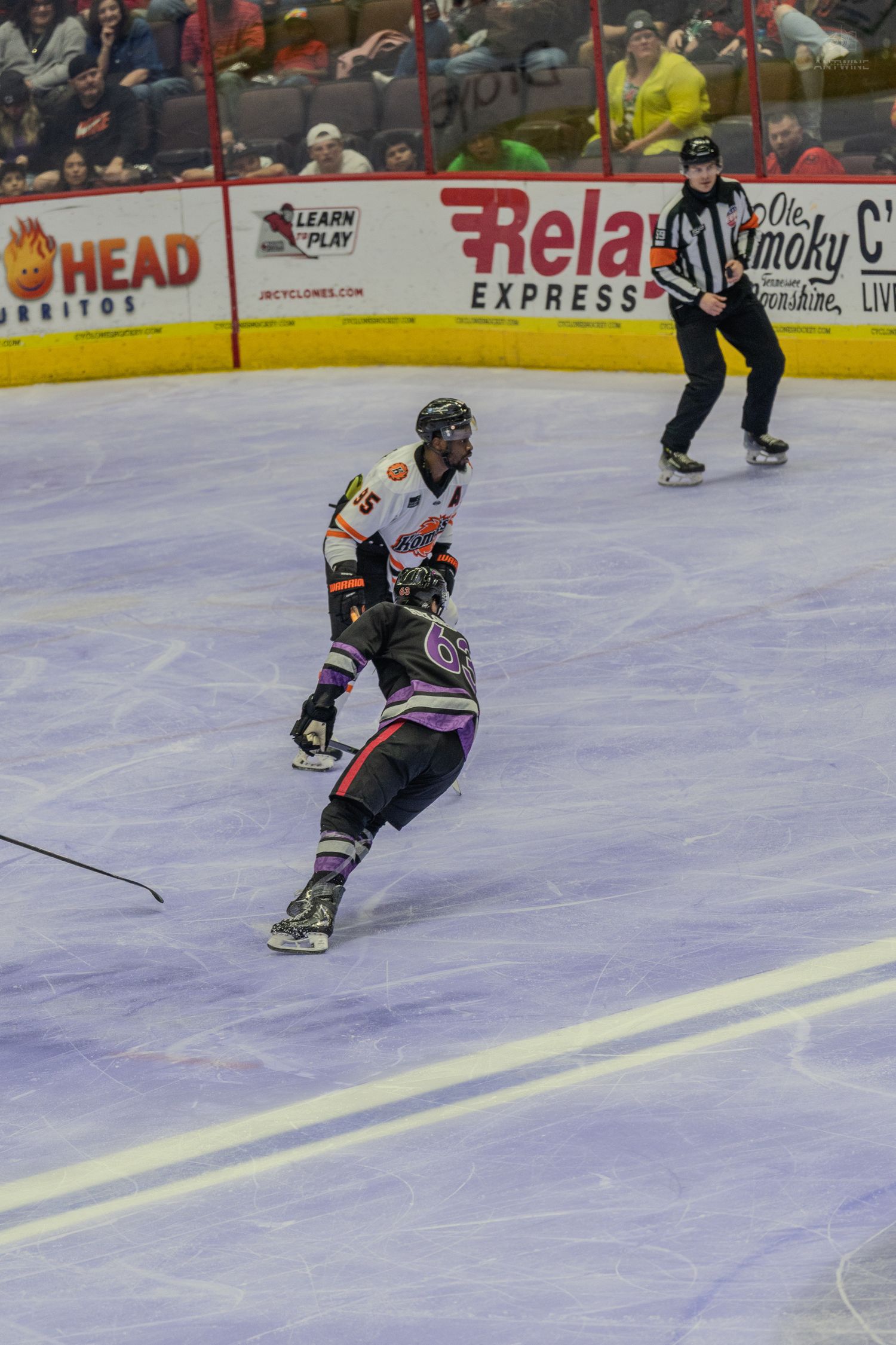 Hockey player in mixed purple and white uniform skates on ice as a referee watches during a minor league game.
