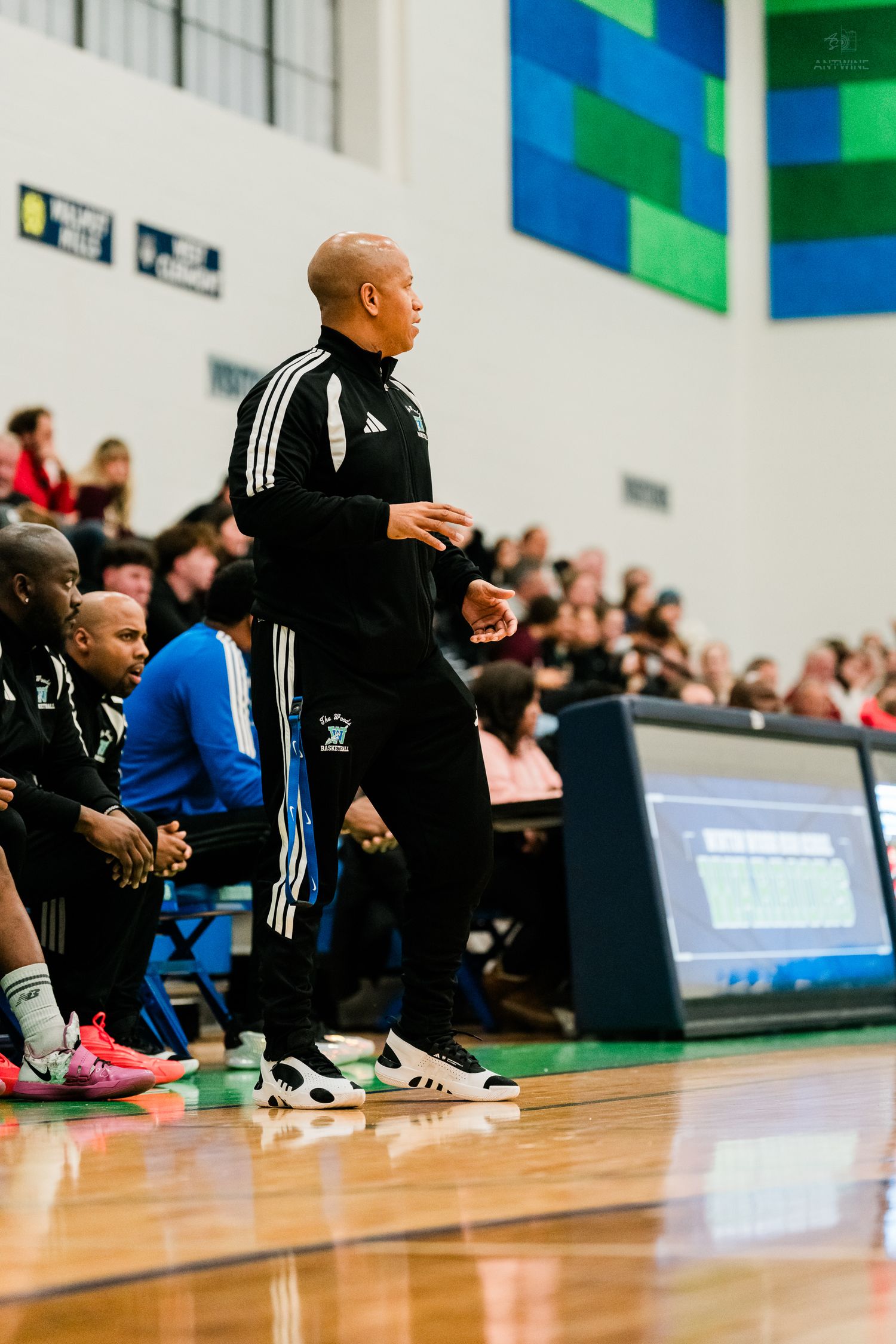 A basketball coach in black Adidas tracksuit stands courtside watching his team during a high school game.