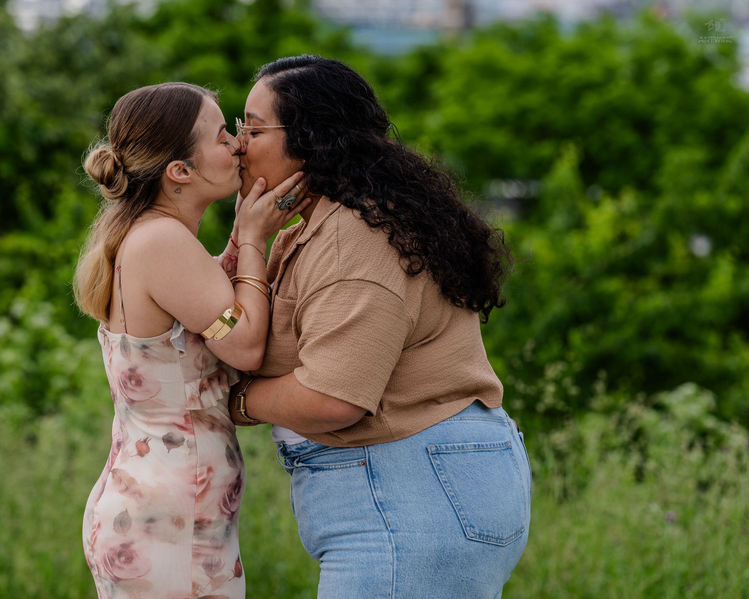 Two women sharing a romantic kiss in a lush green outdoor setting.