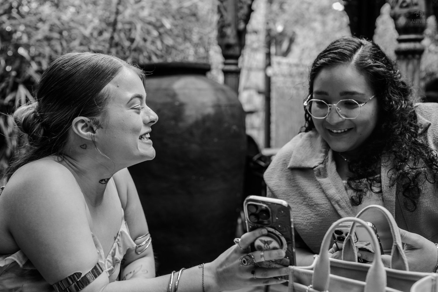 Black and white photo of two laughing women sharing a phone at outdoor cafe.