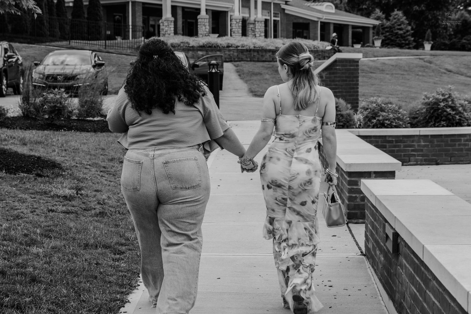 Black and white photo of two women holding hands walking on a sidewalk near a building.