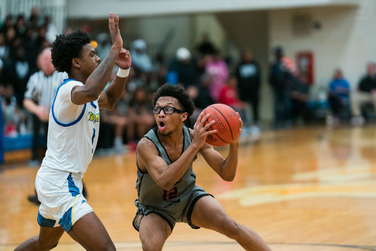 Basketball player in gray drives to basket while defender in blue and white uniform contests the shot.