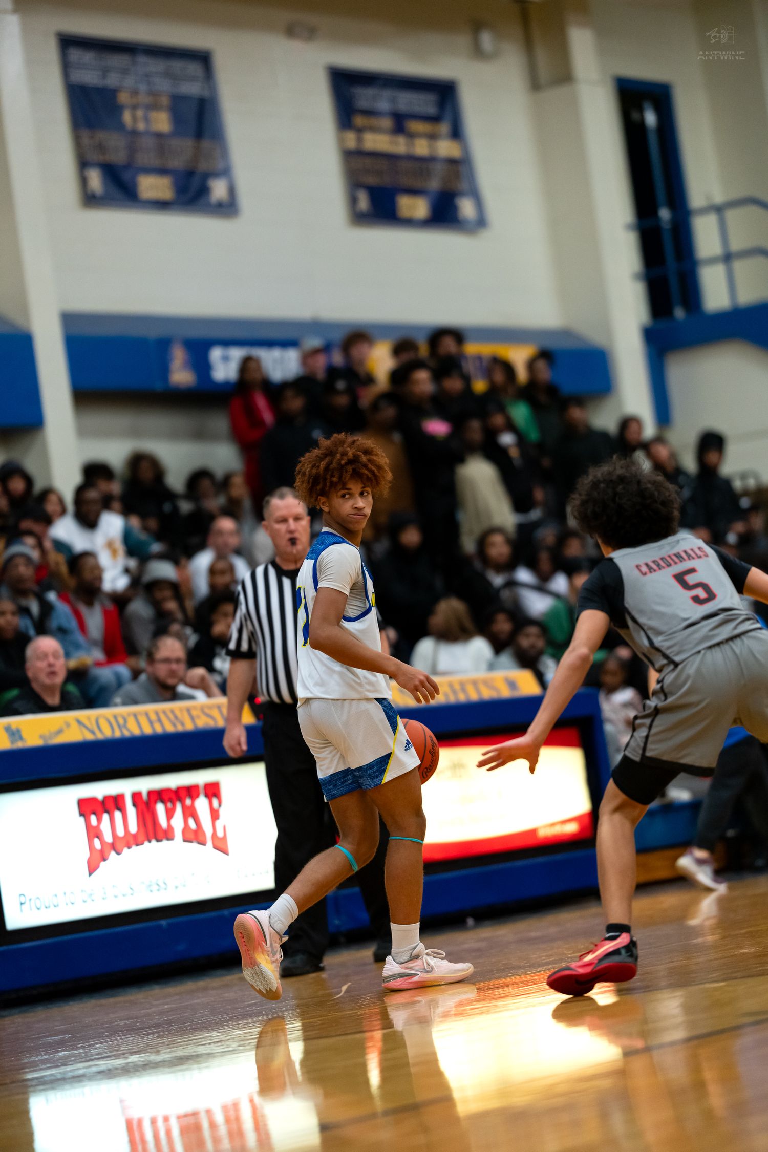 A curly-haired basketball player in white dribbles against a Cardinals defender on the court.