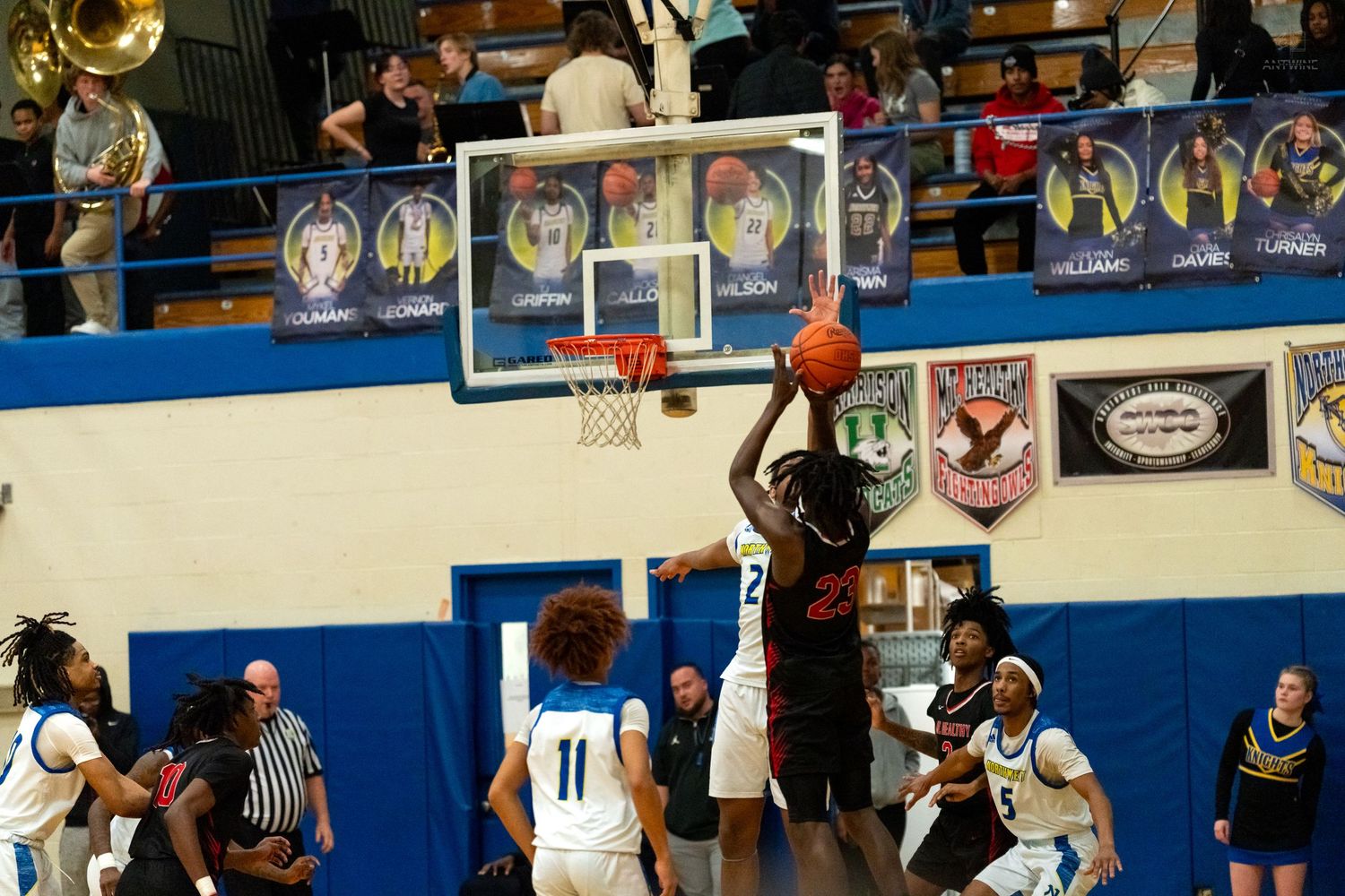 High school basketball player in red attempts a layup over defenders in a packed gymnasium.