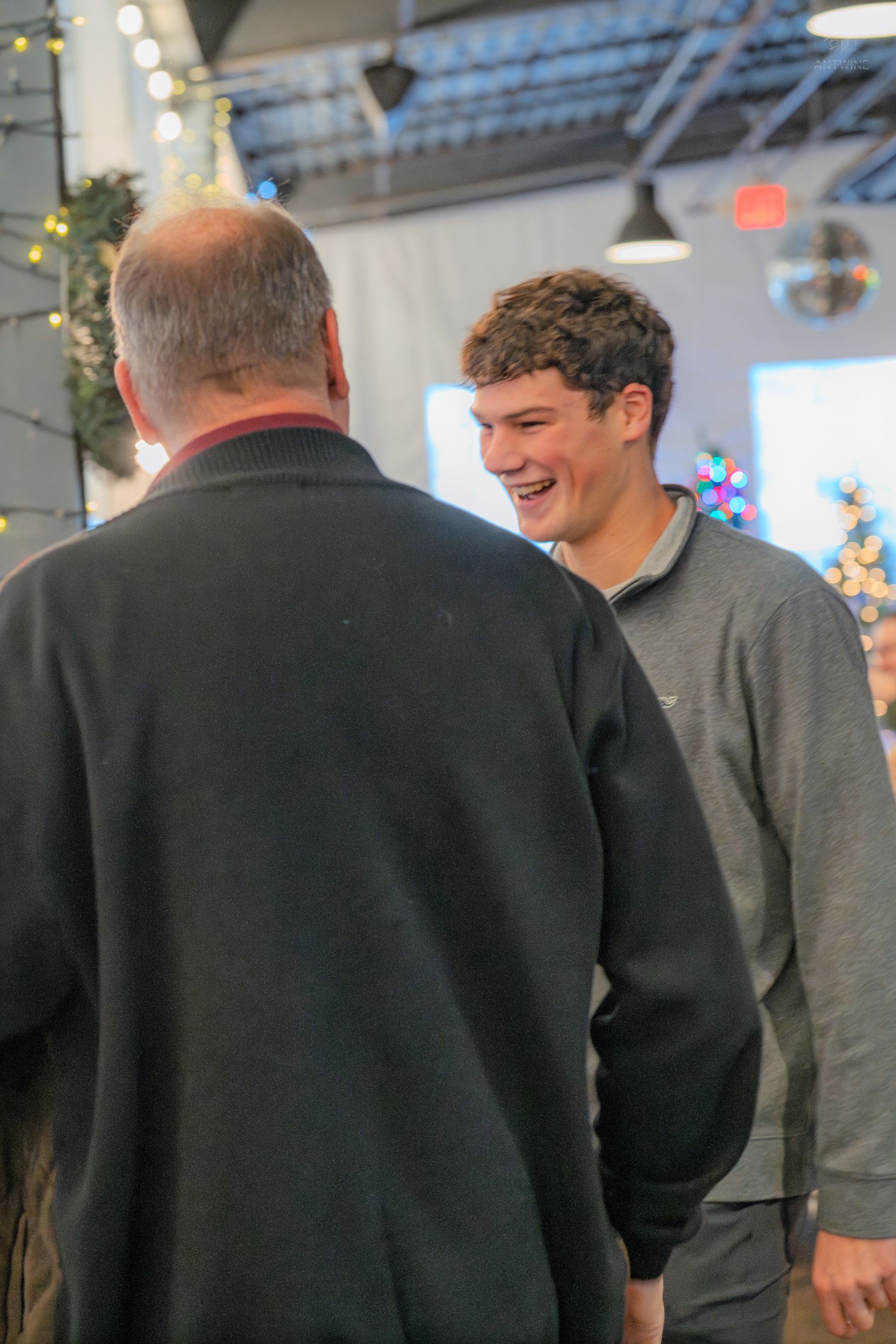 A young man in a gray sweatshirt laughs warmly while conversing with an older man at a festive event.