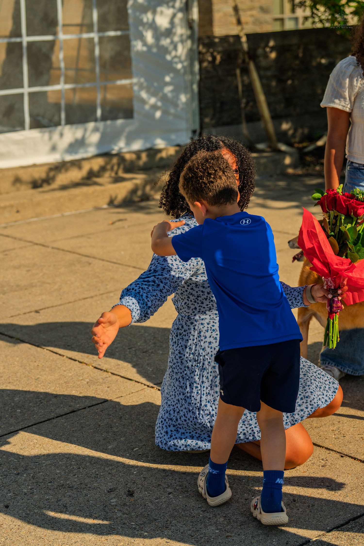 Young boy in blue shirt hugs a woman in a floral dress outdoors while she holds a bouquet of red roses.