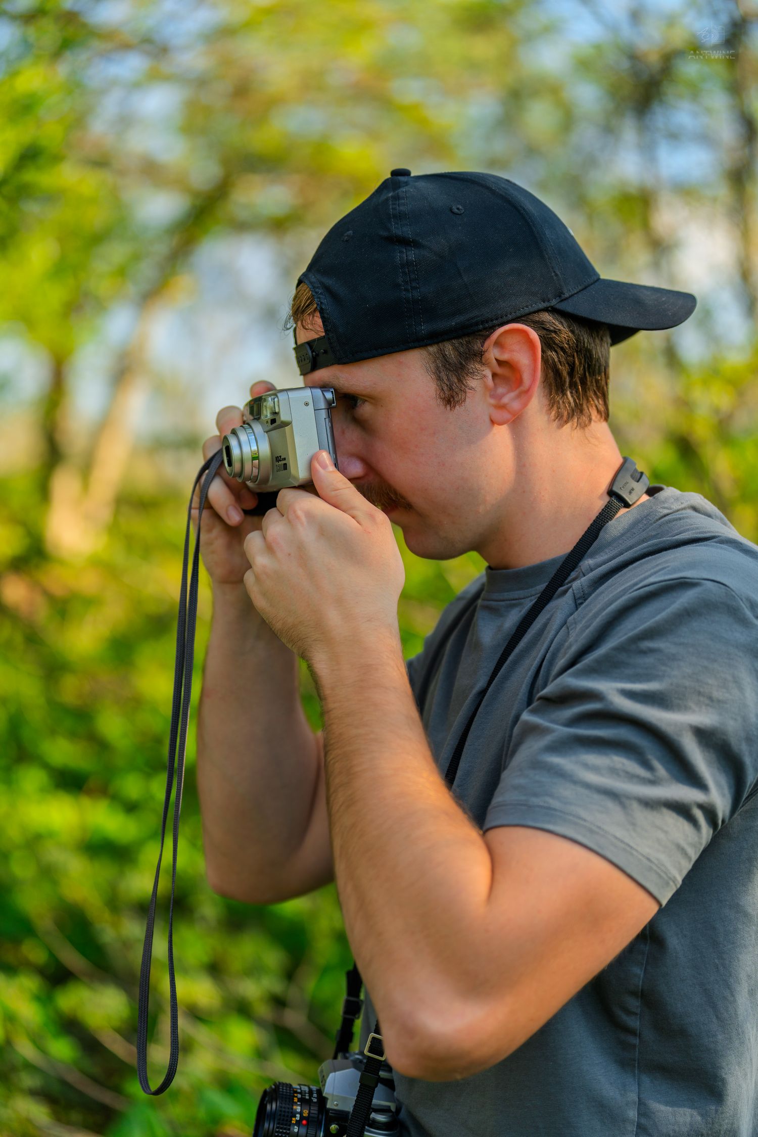 Young man in black cap photographing with vintage film camera outdoors in green nature setting.