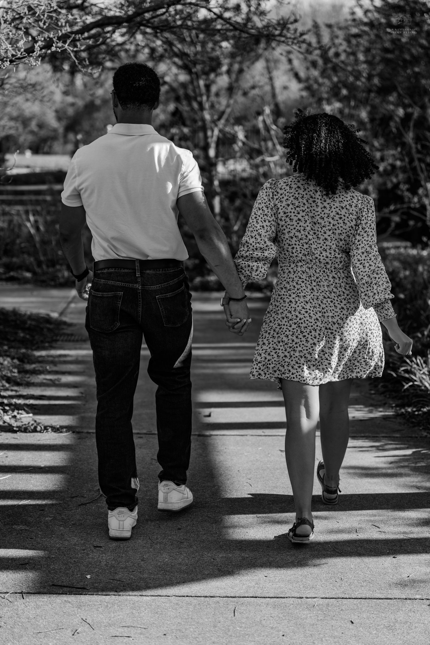 Black and white rear view of a couple holding hands walking along a tree-lined park pathway.