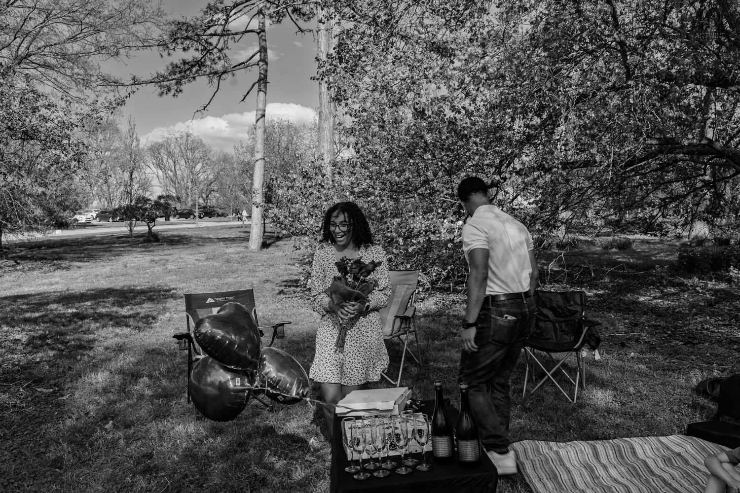 Black and white photo of a couple in a park setting with picnic chairs, balloons, and a bouquet of flowers.
