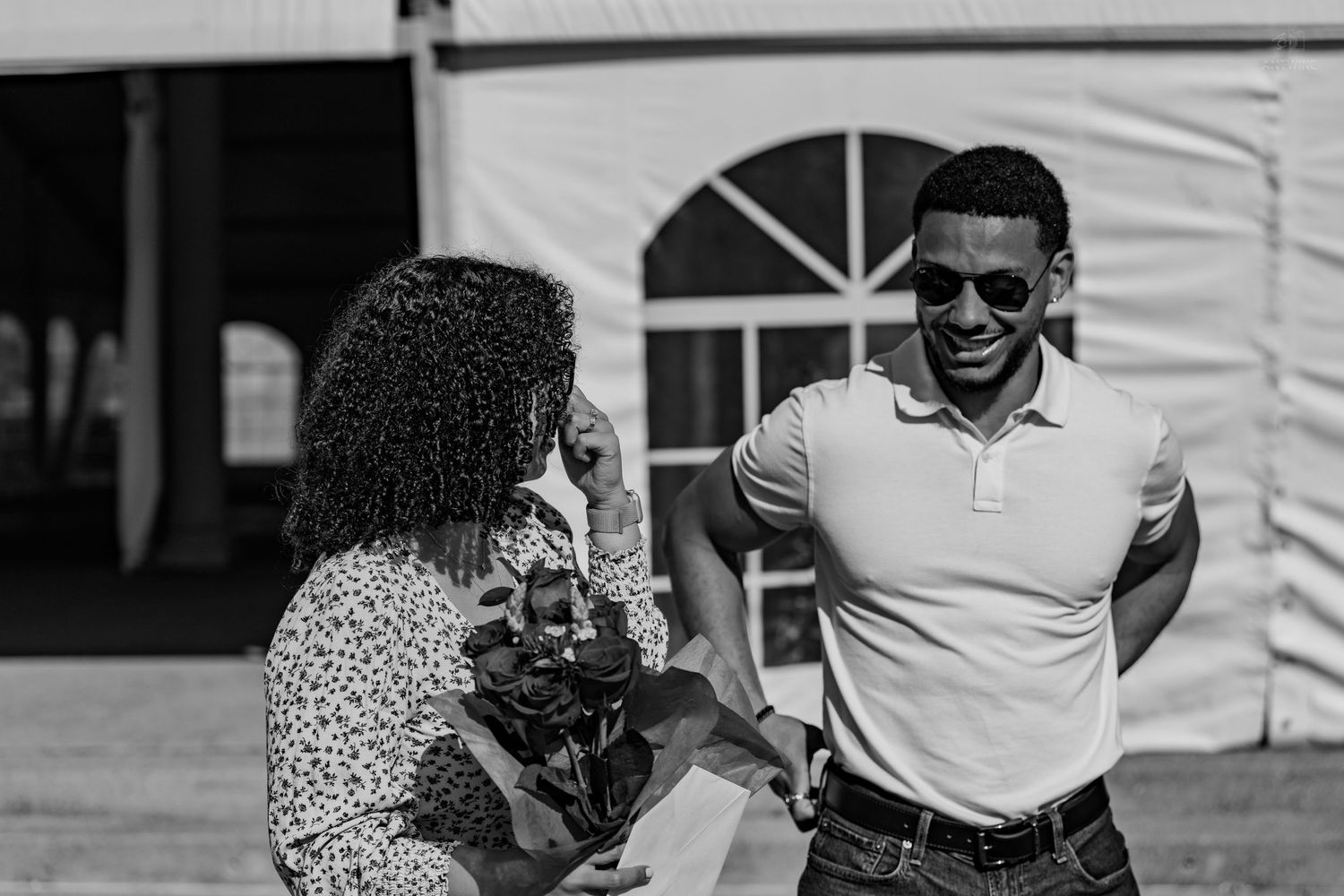 Black and white photo of a laughing couple outdoors, woman holding flowers and man in sunglasses and white polo.