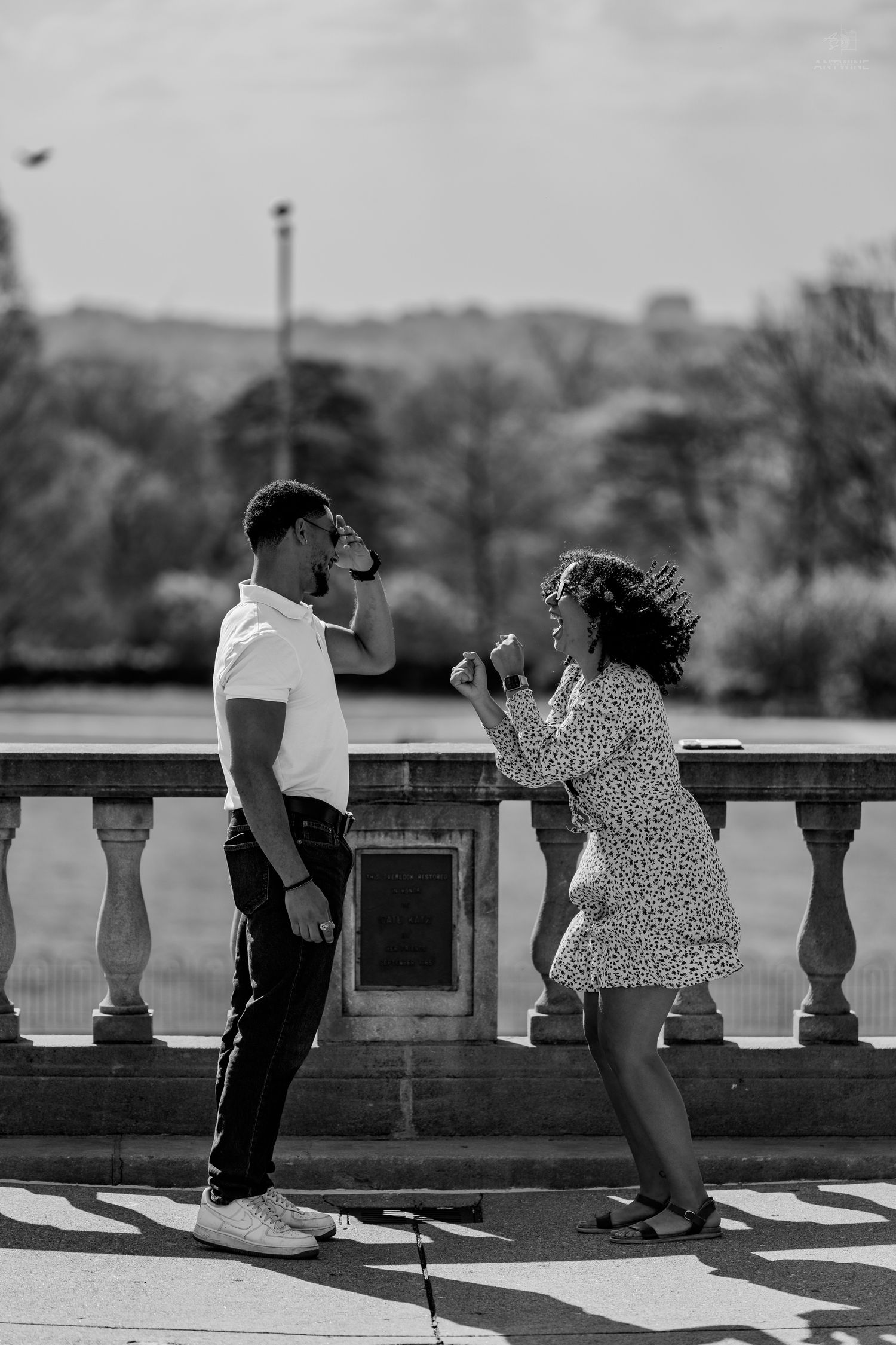 Black and white image of joyful couple celebrating engagement by ornate stone railing in park.