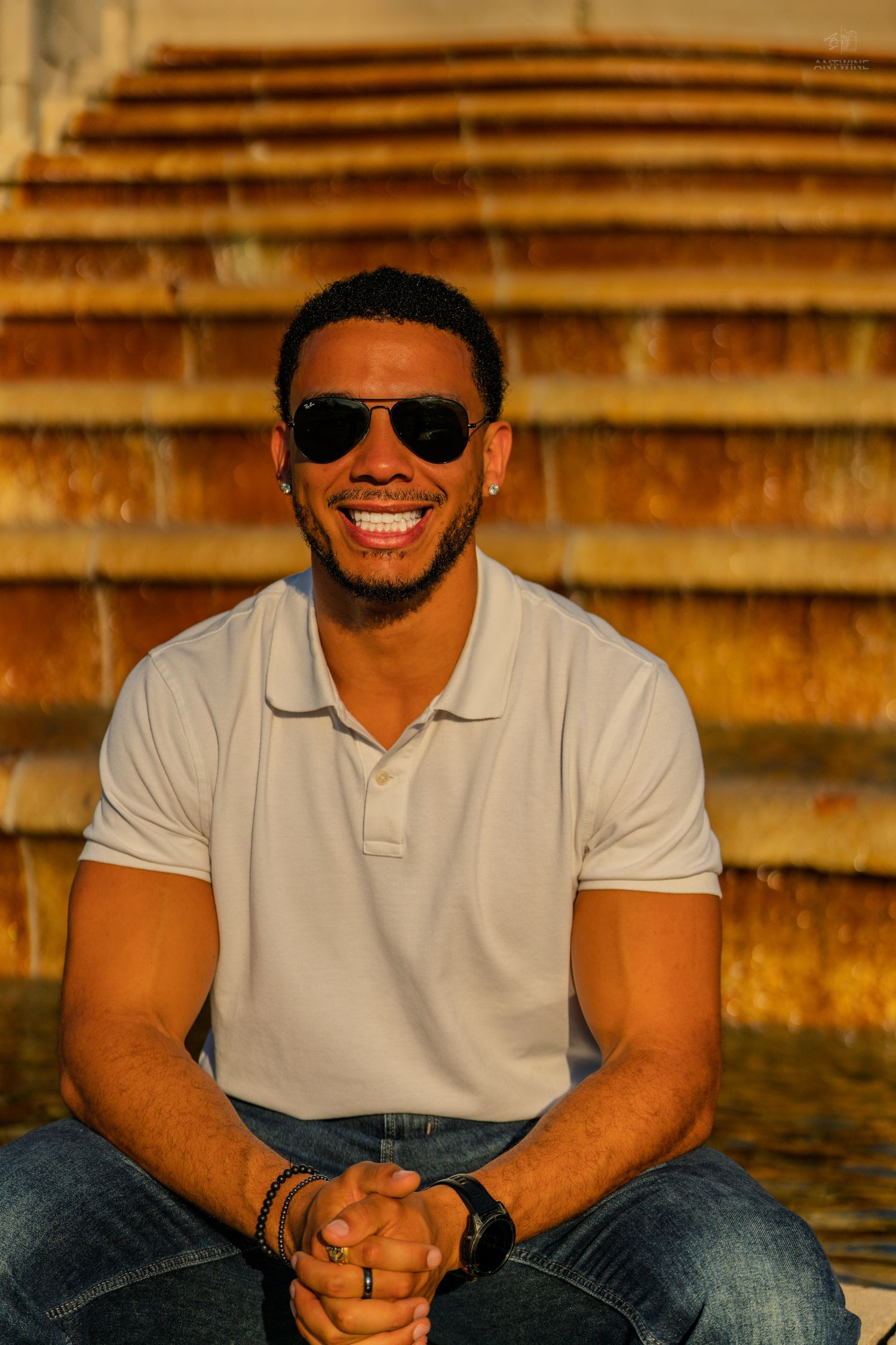 Young man in white polo shirt and aviator sunglasses smiling while sitting on golden-lit stone steps.