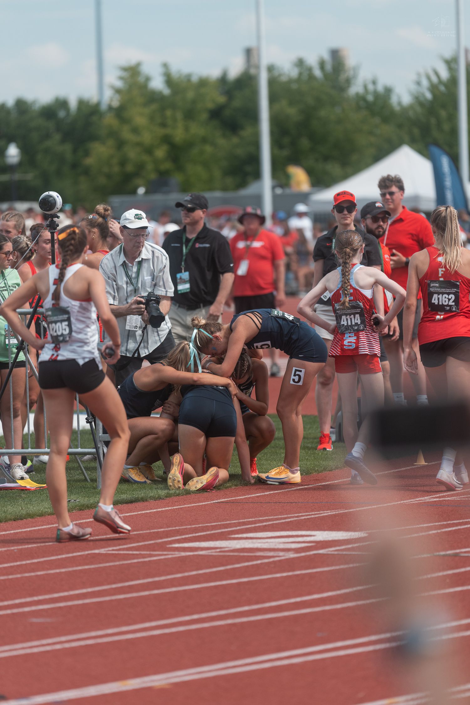 Female track athletes and photographers crowd near the finish area after completing a relay race event outdoors.