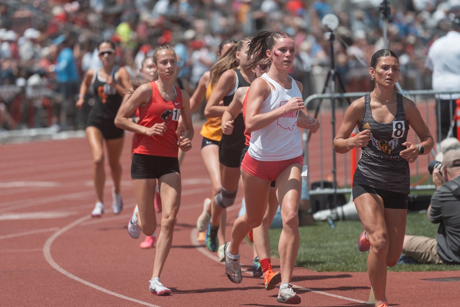 Female high school runners compete in a distance track race before a large crowd at a sunny outdoor meet.