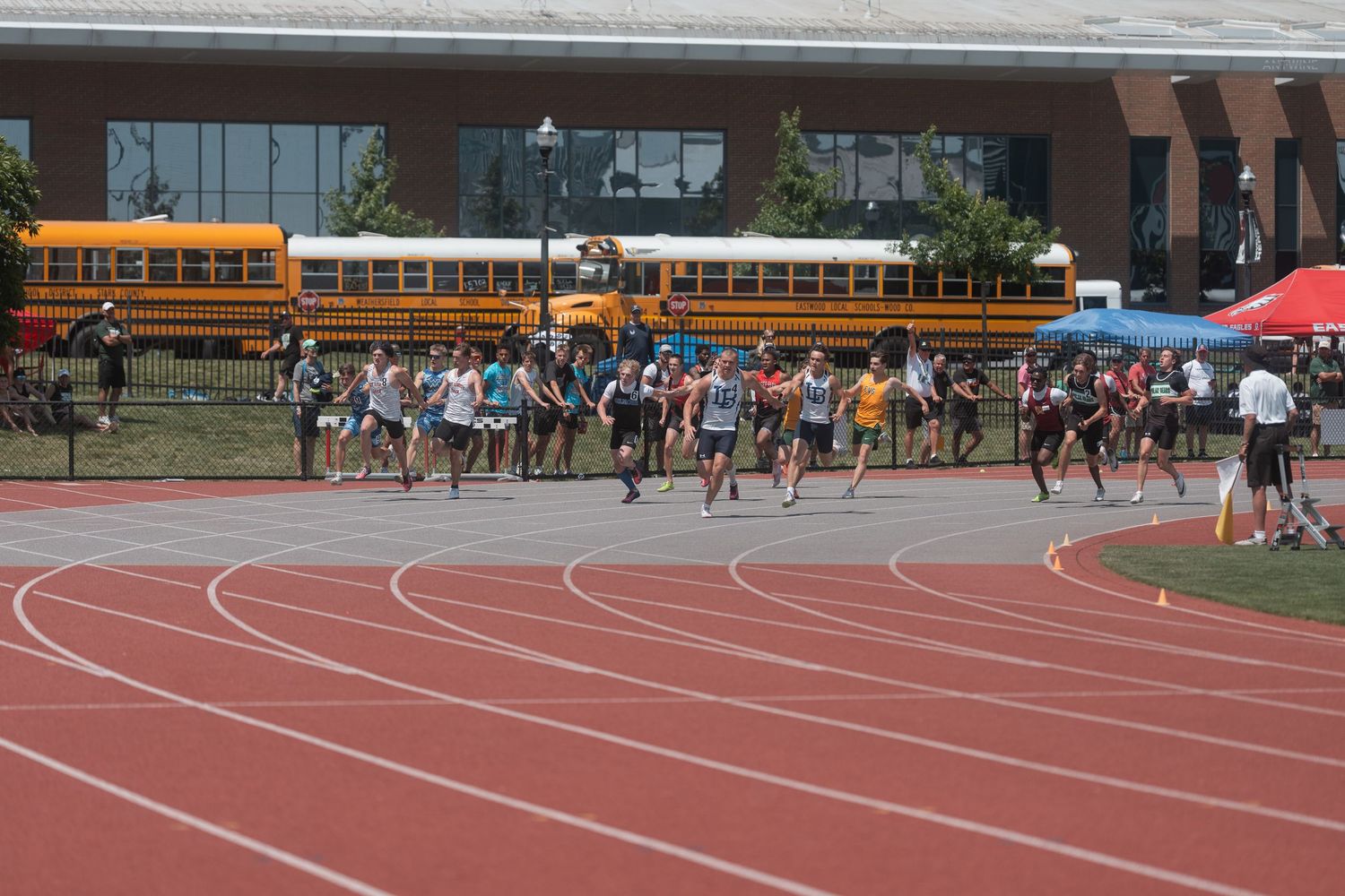 A large group of runners compete in a distance race on a red outdoor track at a championship meet.