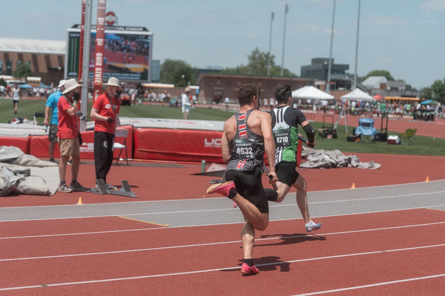 Two runners compete in a relay race on a red track at a sunny outdoor athletic championship.