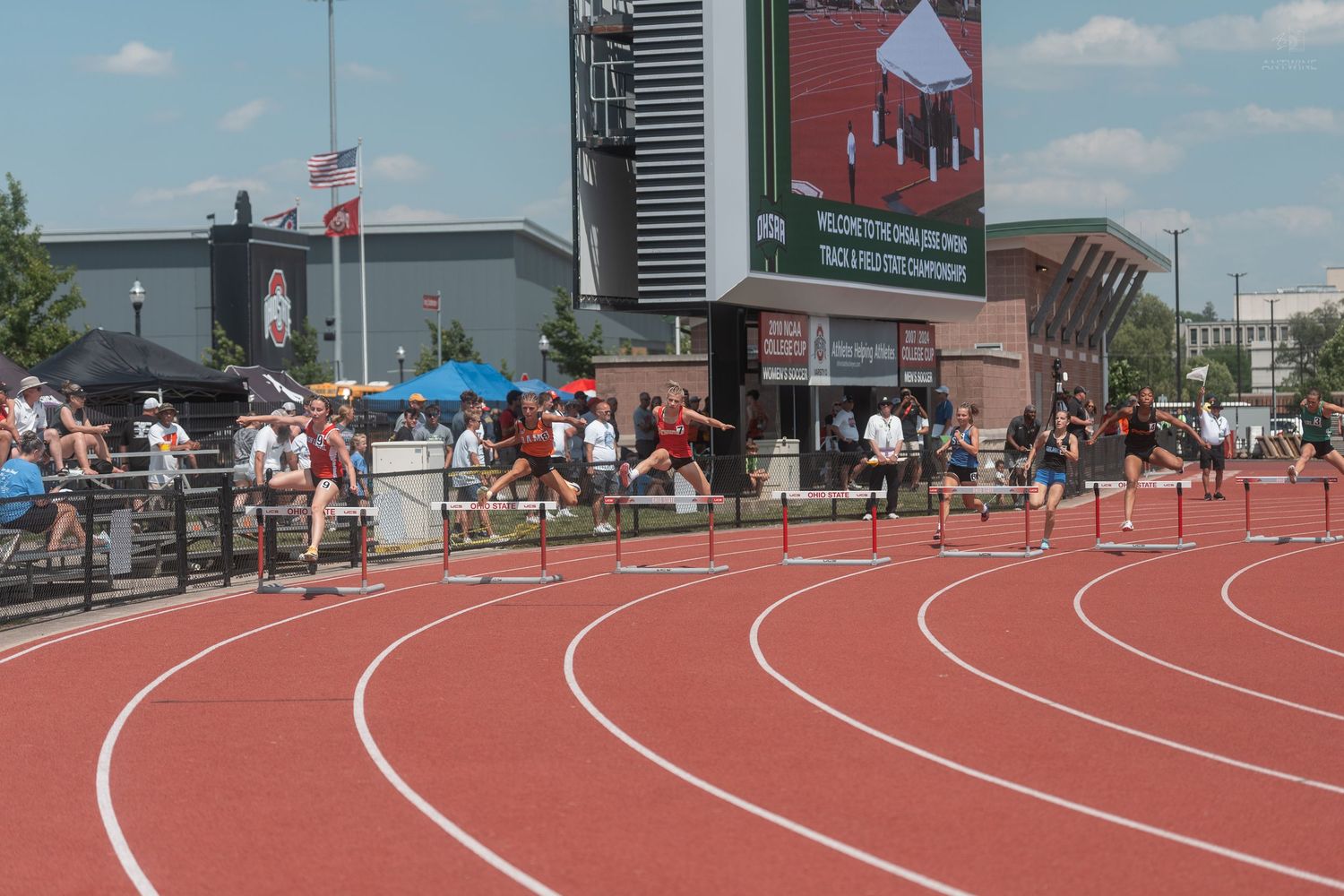 Athletes clearing hurdles on a red outdoor track at a competitive meet with a large stadium scoreboard in background.