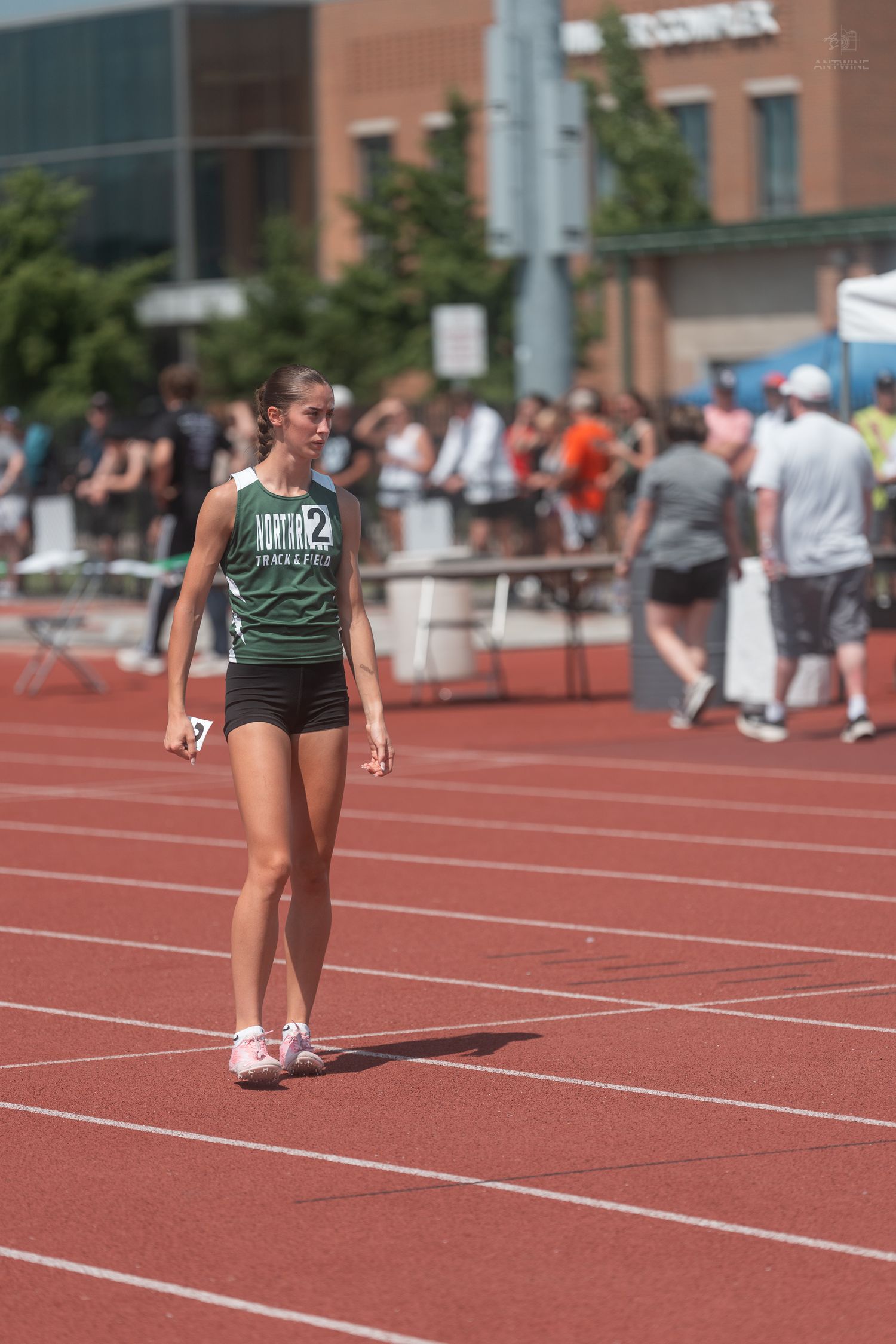 Young female runner in green Mantis track uniform walks on red track during sunny outdoor athletics meet.