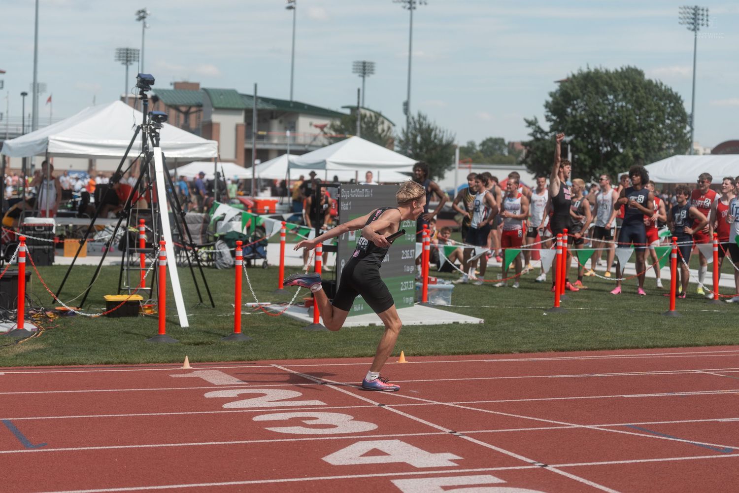 Athlete crosses finish line at outdoor track and field meet with crowd of spectators cheering in background.