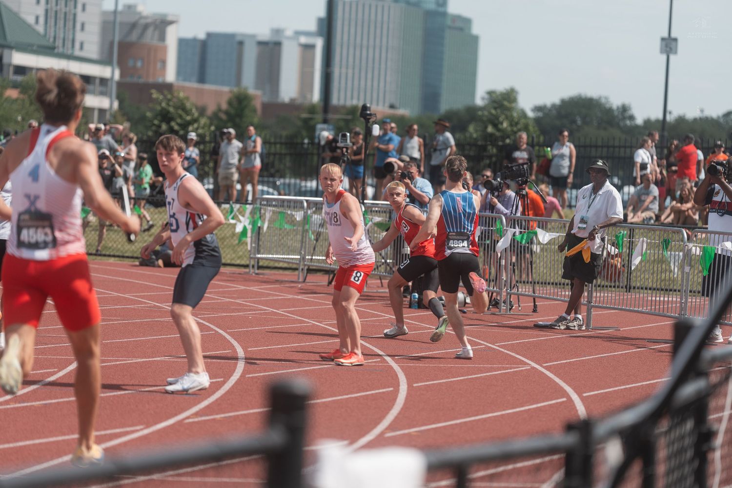 Male track athletes pass baton during competitive outdoor relay race with spectators watching.