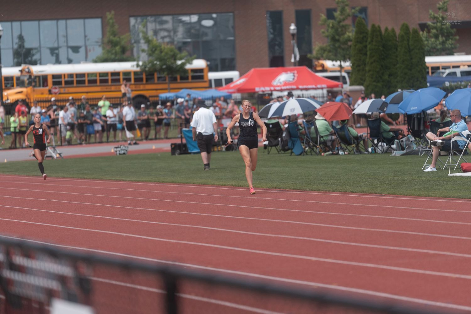 Female runners compete on a red track at an outdoor high school track and field competition.