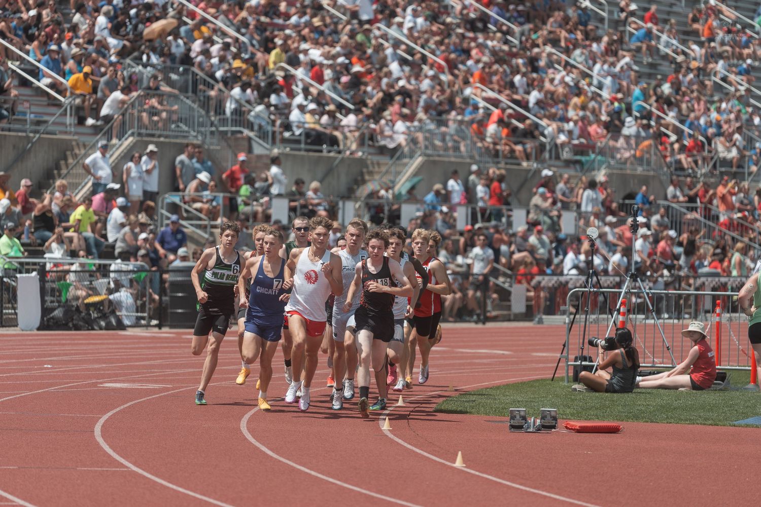 Male runners compete in a distance race on a red track at a well-attended high school track and field meet.